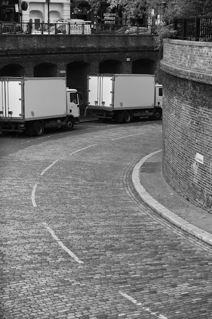 A curved cobblestone street in an urban area featuring two white box trucks parked side by side, with the rear sections visible. The trucks are positioned near a brick retaining wall and a building wi